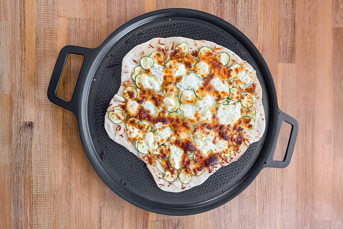 A freshly baked pizza topped with zucchini slices, melted cheese, and herbs sits on a round pizza pan with handles, placed on a wooden table.