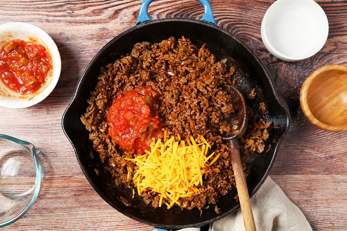 overhead shot of a black cast iron skillet with blue handle on a wooden table, filled with cooked ground beef, onions, salsa, and shredded cheese; a white bowl with salsa on the left side of the skillet; a white bowl with no contents on the right side of the skillet; a glass measuring cup on the left side of the skillet; a wooden bowl on the right side of the skillet; a brown cloth beneath the skillet on the right side of the image and wooden spoon in the skillet