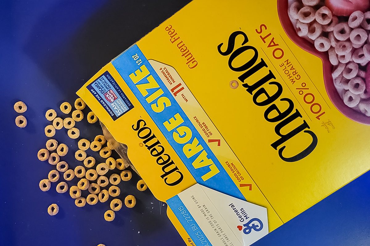 A large box of Cheerios cereal is tipped over, spilling circular cereal pieces onto a blue surface. The packaging highlights that it