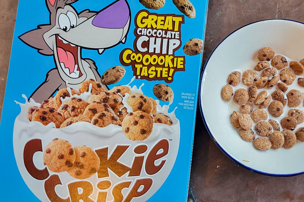 Box of Cookie Crisp cereal with a cartoon wolf and a bowl filled with chocolate chip cookie-shaped cereal and milk. The box features the text 