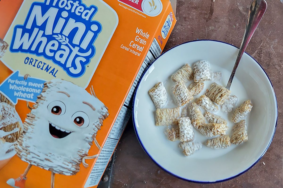 A bowl of Frosted Mini-Wheats with milk and a spoon sits beside an open box of the cereal on a countertop. The cereal box displays a smiling wheat character.