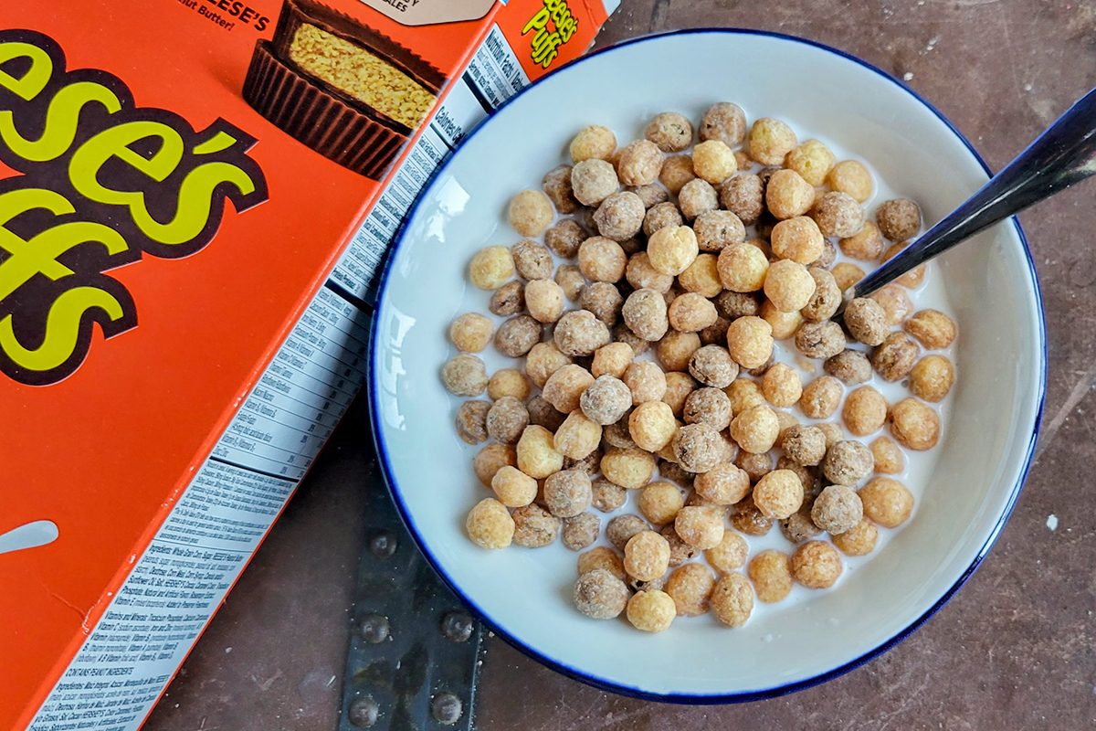 A bowl of cereal with milk and a spoon, featuring spherical, crunchy pieces. A partial view of a Reese