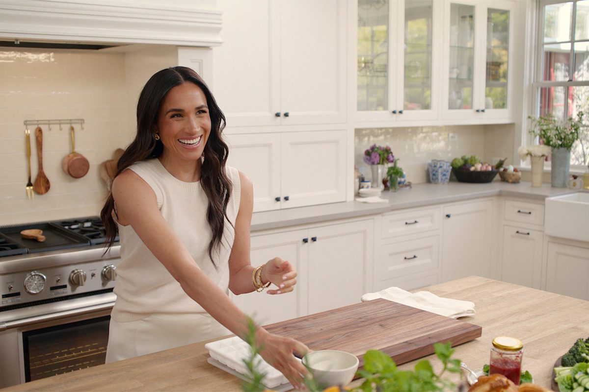 Meghan Markle in a white dress stands in a bright kitchen, smiling as she prepares food on a wooden countertop. The kitchen has white cabinets, a stove, and various kitchen items.
