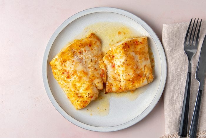 Two golden-brown baked fish fillets on a white plate. The fillets are drizzled with a light sauce. A fork and knife are placed on a beige napkin to the right of the plate. The background is a soft pinkish hue.