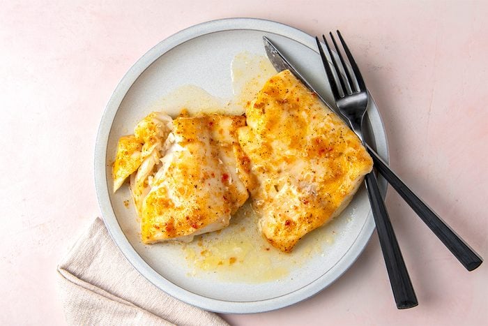 Two pieces of cooked white fish topped with a golden sauce are served on a round white plate. A fork and knife rest beside the fish. The plate is placed on a light pink surface next to a folded beige napkin.