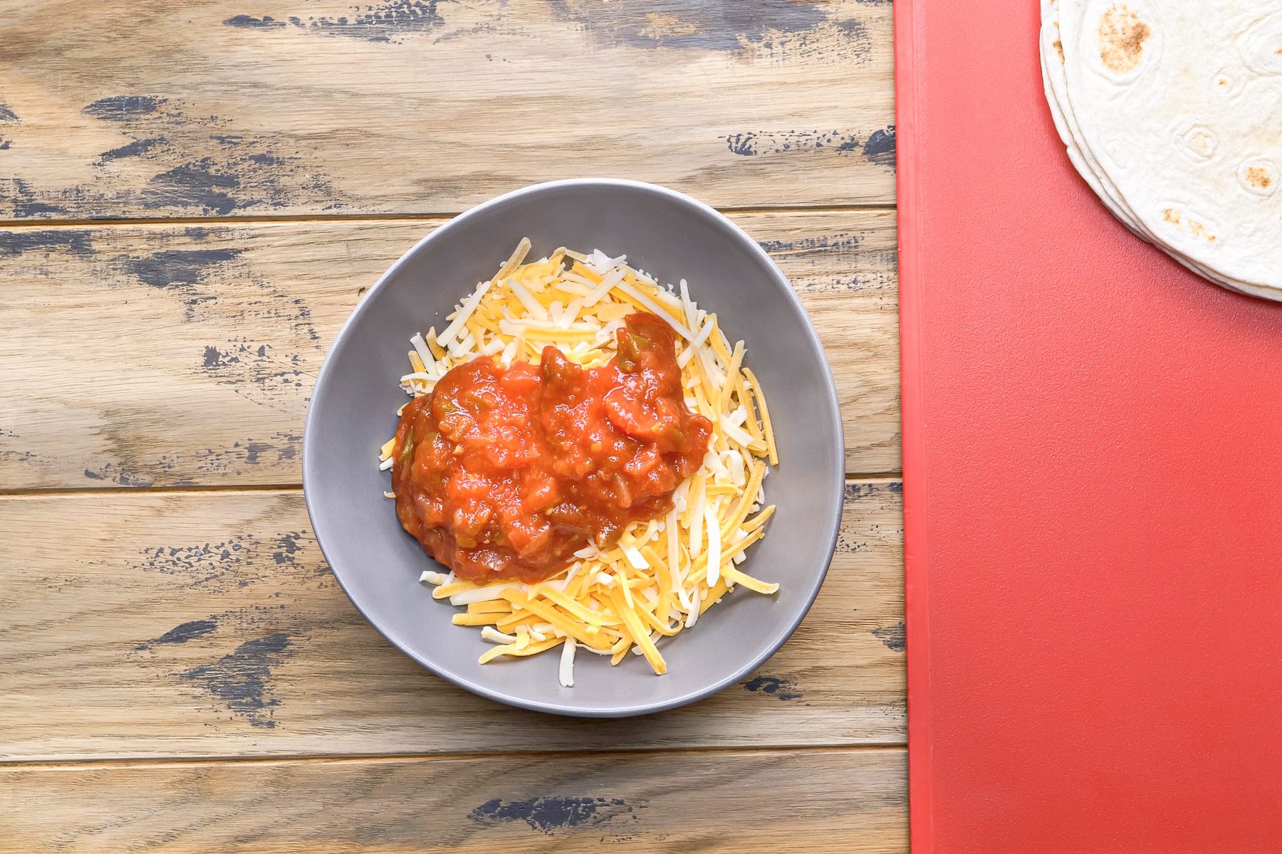 overhead shot of a bowl of shredded cheese and salsa sitting on a wooden table; next to the bowl on the right is a red cutting board with a stack of tortillas on it