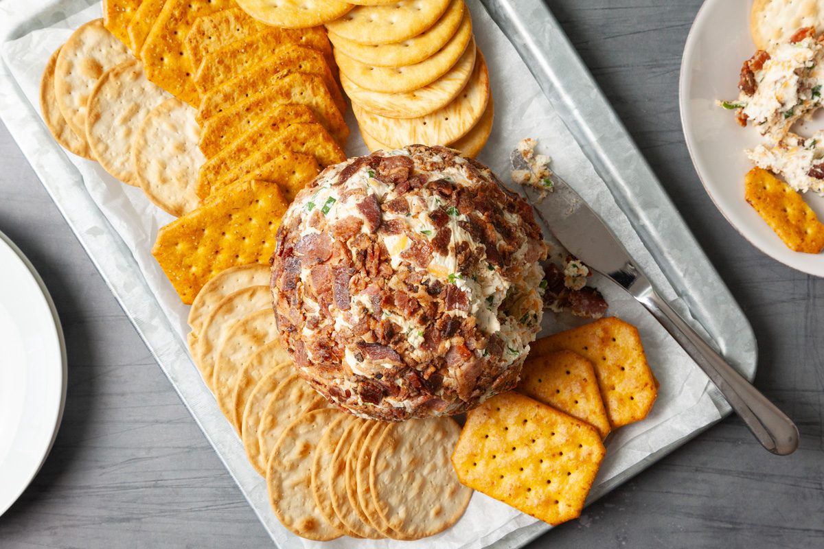 Overhead shot of Bacon Ranch Cheese Ball; on a platter with assorted crackers; served on plate; knife; empty plates; grey surface