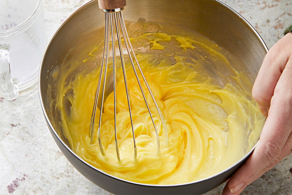 3/4th shot of a mixing bowl filled with a creamy yellow batter, A stainless steel whisk is immersed in the mixture, To the left of the bowl, there is a clear glass filled with water; The background consists of a light colored surface