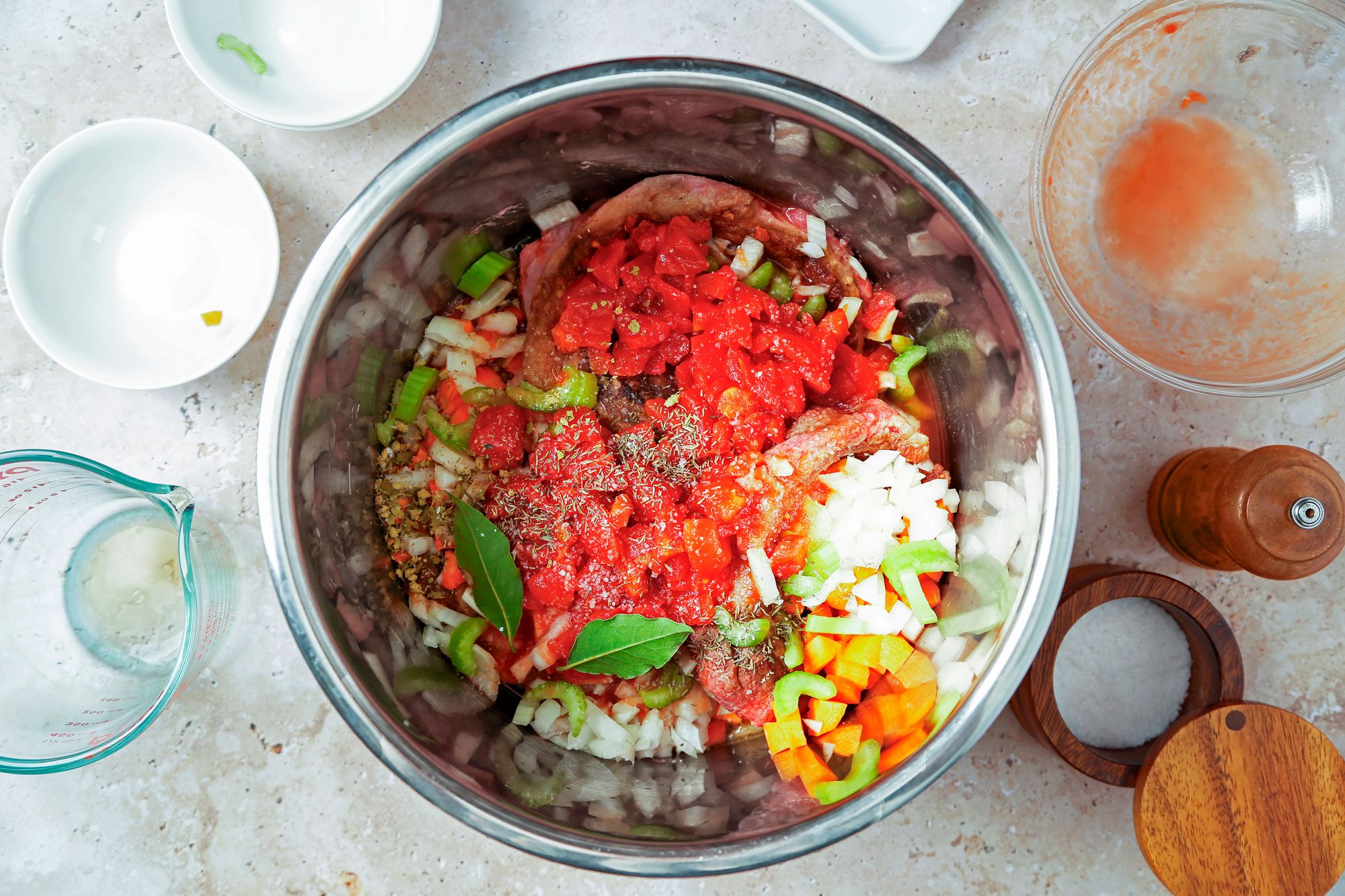 overhead shot of the ingredients for a recipe, likely a stew or soup, arranged on a light beige countertop; a large stainless steel bowl sits in the center of the image, the bowl sits on a beige countertop next to other ingredients like a small glass measuring cup, a small white bowl, a glass bowl with a reddish-brown tint, a wooden salt shaker, and a wooden pepper grinder