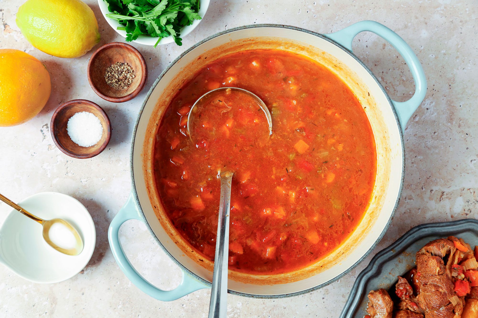 overhead shot of a pot of soup with a ladle inside, a bowl of parsley, two bowls with salt and pepper, a plate of meat and vegetables, and a bowl with a spoon