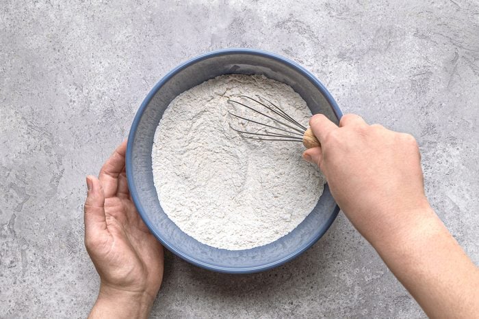overhead shot of a pair of hands holding a blue bowl filled with flour or dry baking ingredients, a whisk with a wooden handle is inside the bowl, partially submerged in the powder; the background is a textured grey surface