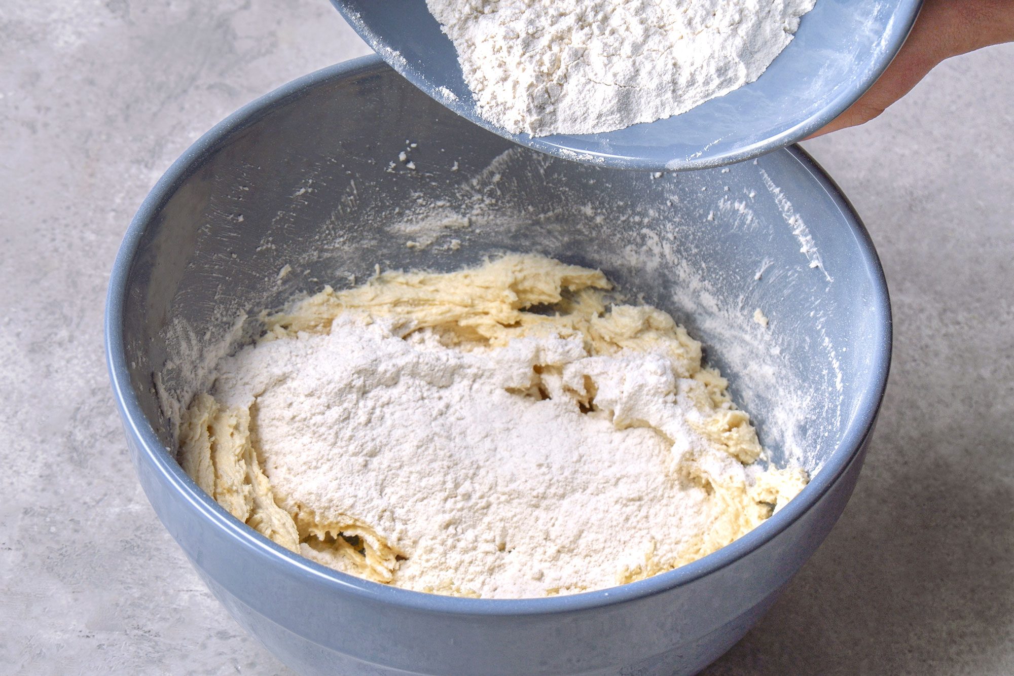 3/4th shot of hands pouring white flour from a light blue bowl into a larger, the background is a grey