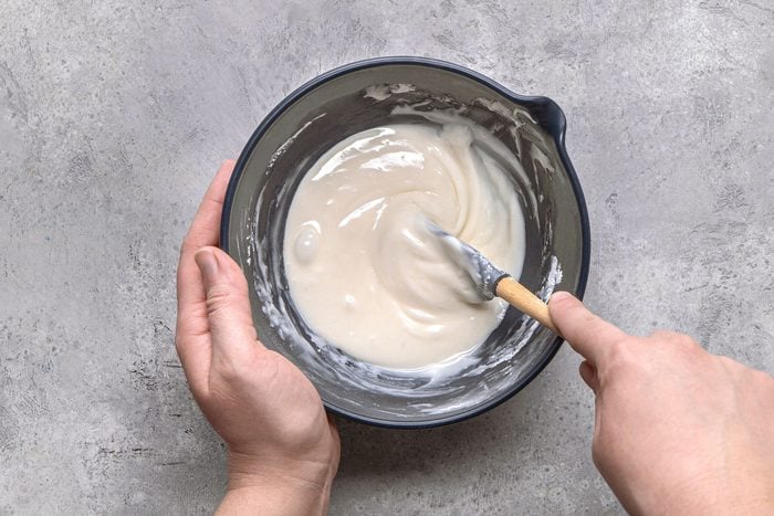overhead shot of a pair of hands holding a blue bowl, inside which a smooth, white glaze is being stirred with a spatula, and the background is a textured grey surface