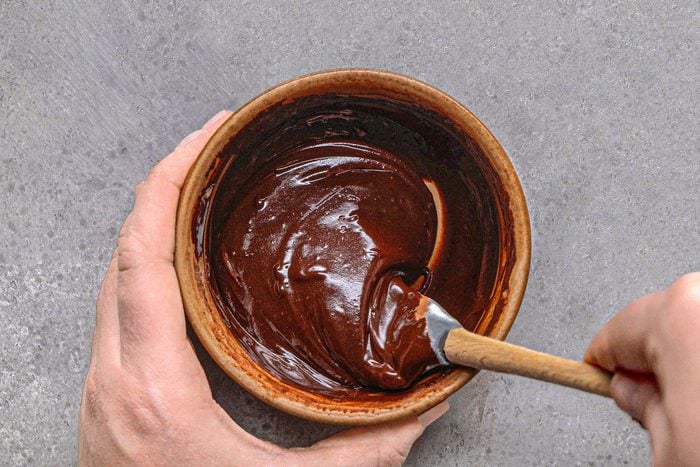 overhead shot of a pair of hands holding a brown bowl, inside which a chocolate ganache, is being stirred with a spatula, and sits on a textured grey surface