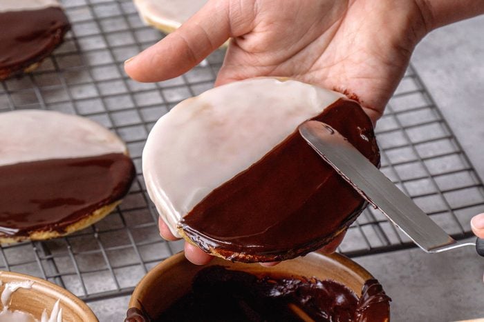 A close-up shot of hands using a spatula to apply dark chocolate frosting to a round cookie, a bowl containing more dark chocolate frosting is visible below, and several already frosted black and white cookies are in the blurred background on a wire rack, the background is a textured grey surface