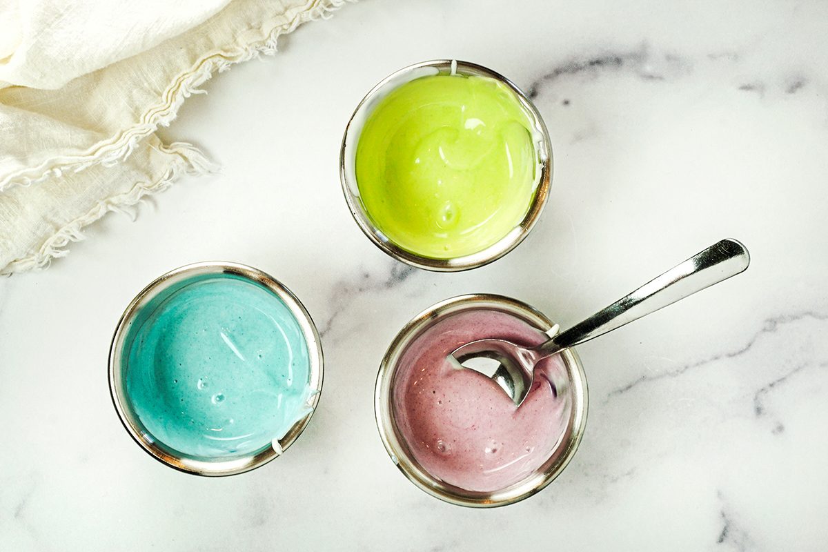 Three bowls of colorful whipped cream on a marble surface. The bowls contain pastel green, blue, and purple cream. A spoon rests in the purple bowl, and a piece of white fabric is in the upper left corner.