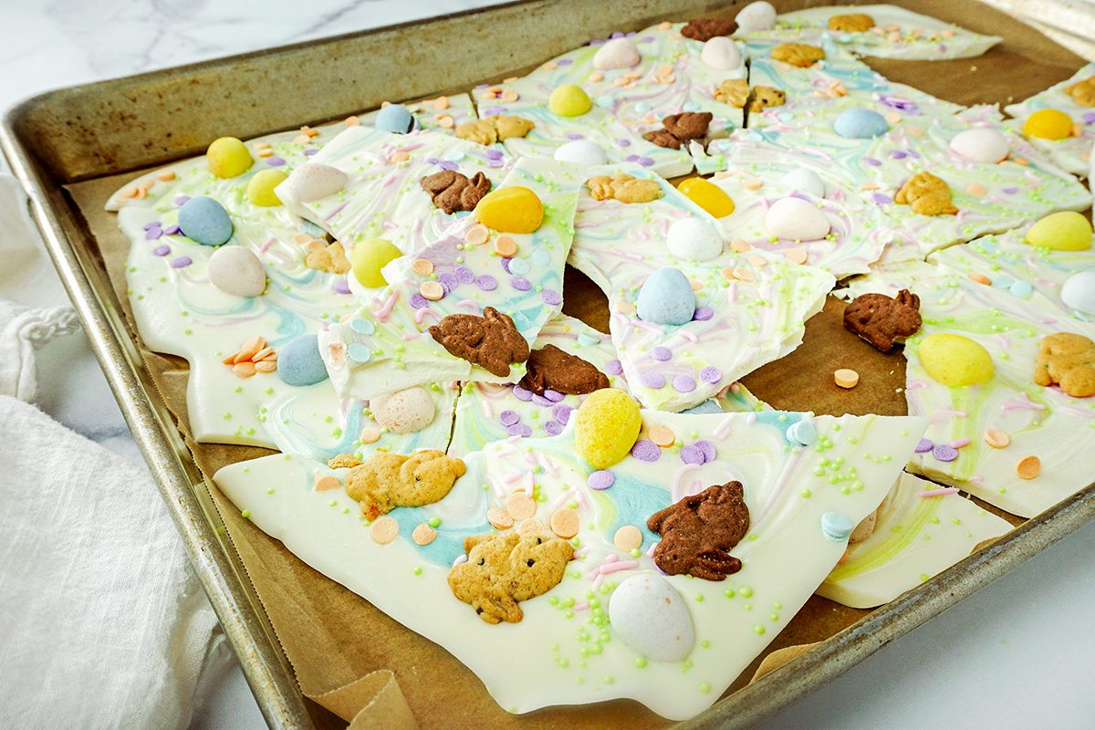 A baking sheet filled with festive white chocolate bark topped with colorful candy eggs, pastel sprinkles, and small chocolate bunnies. A white cloth is partially visible on the side.