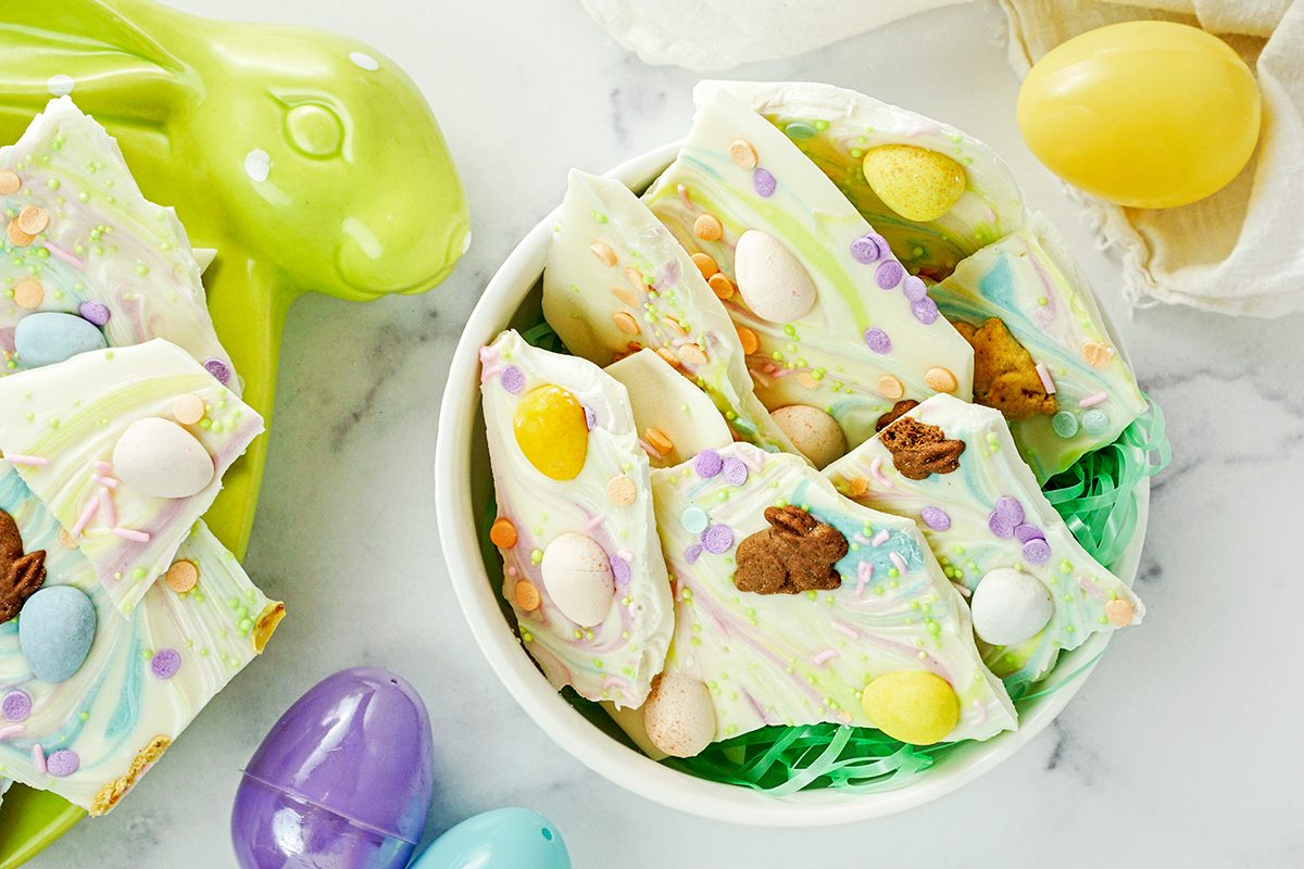 A bowl filled with colorful Easter-themed chocolate bark pieces, topped with pastel candies and decor. Surrounding the bowl are plastic Easter eggs, and a green rabbit-shaped dish on a marble surface.