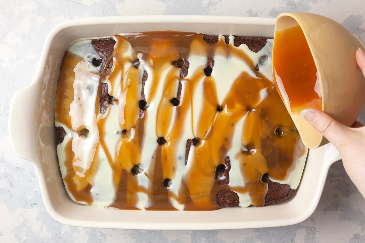 A hand is pouring caramel sauce from a brown bowl onto a chocolate poke cake in a rectangular white baking dish. The cake is covered with stripes of condensed milk and caramel sauce.
