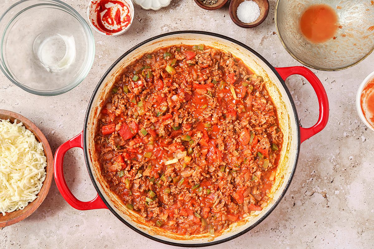 A red pot filled with chunky meat sauce, including visible pieces of tomatoes and green peppers, is placed on a speckled countertop. Surrounding the pot are empty bowls and a dish of shredded cheese.