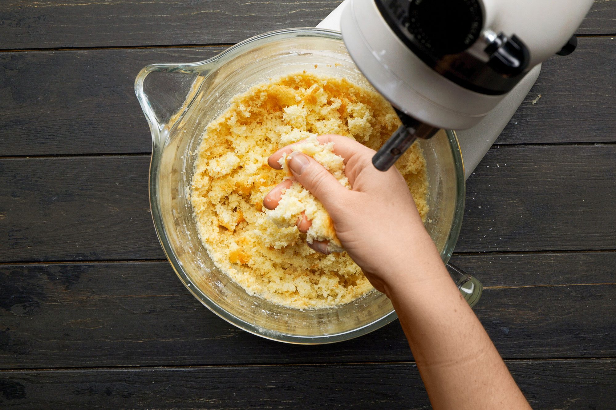overhead shot of a glass mixing bowl filled with a crumbly mixture, likely a cake or cookie batter, a hand is seen gripping a portion of the mixture, the backdrop consists of a dark wooden surface