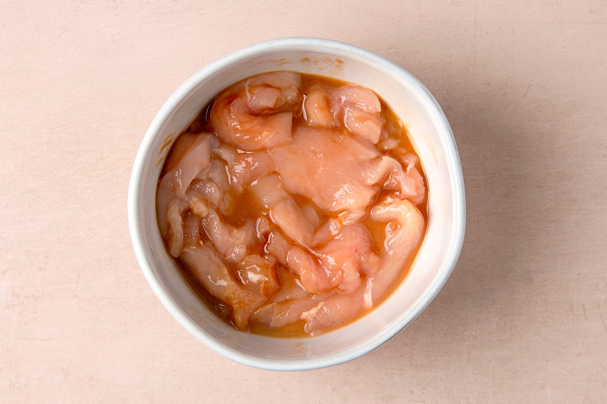 A white bowl filled with raw, marinated chicken pieces on a light wooden surface. The chicken is coated in a light brown sauce.