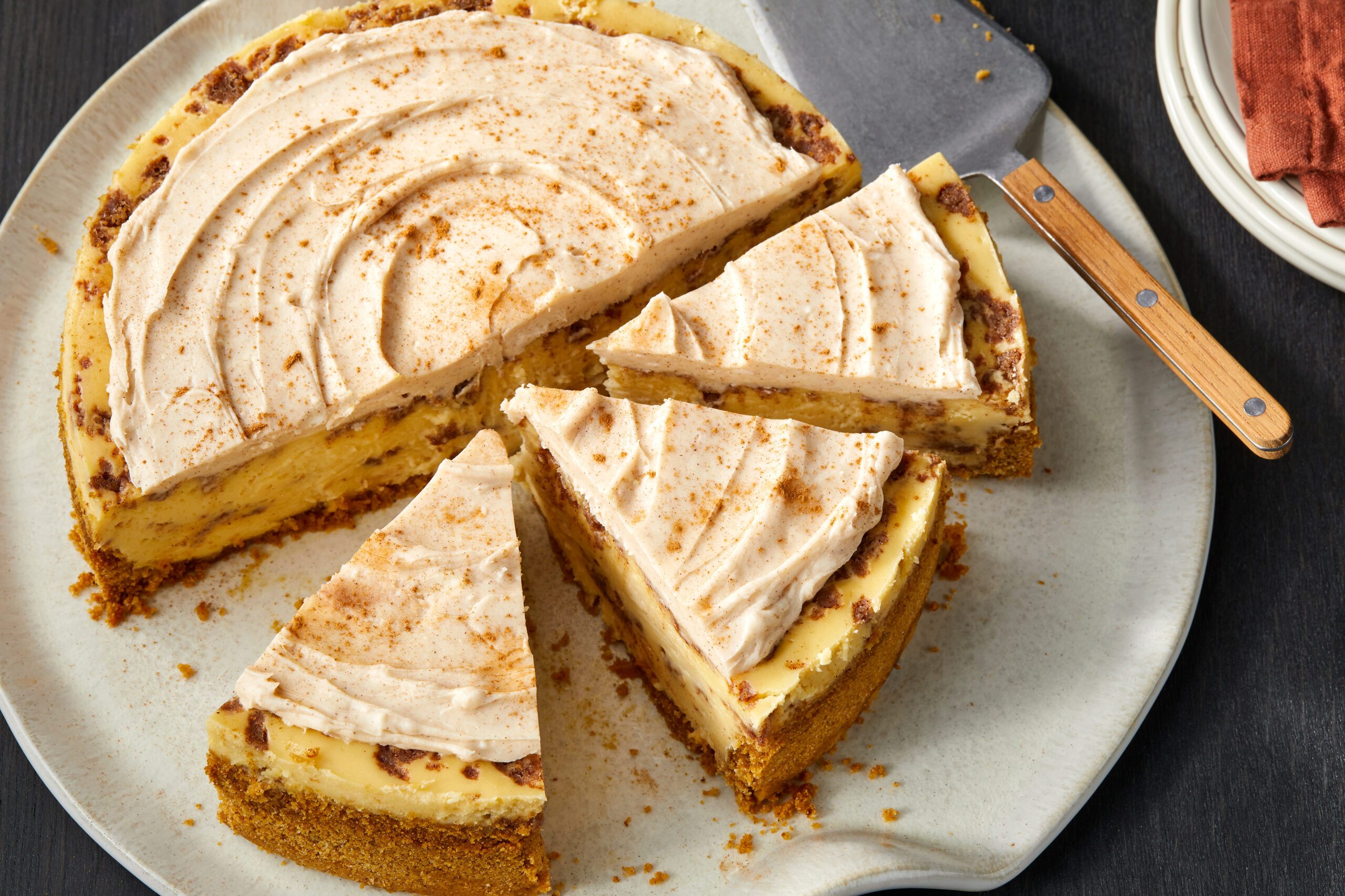 A cheesecake topped with a creamy frosting and dusted with cinnamon, served on a beige plate. Several slices are cut and separated, with a serving spatula nearby. The dark tabletop and some stacked white plates are in the background.