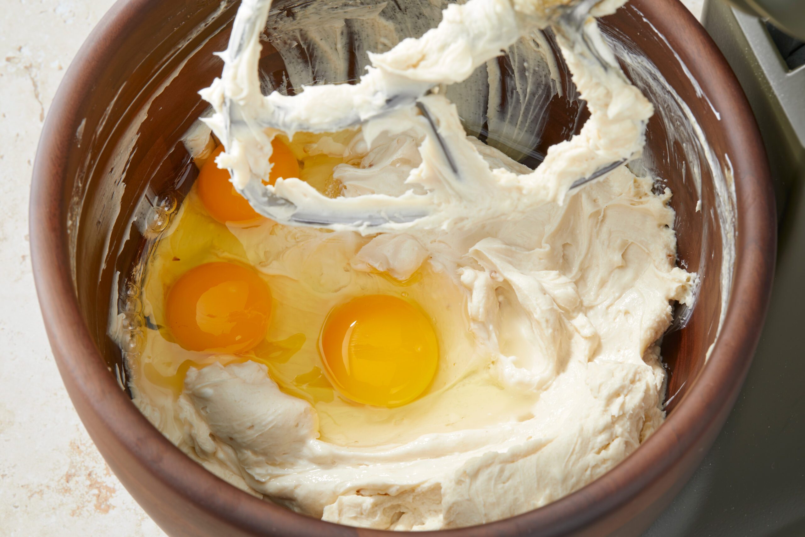 A mixing bowl containing a creamy, beige batter with three cracked eggs on top. The metal beaters are above the mixture, suggesting a baking process in progress.
