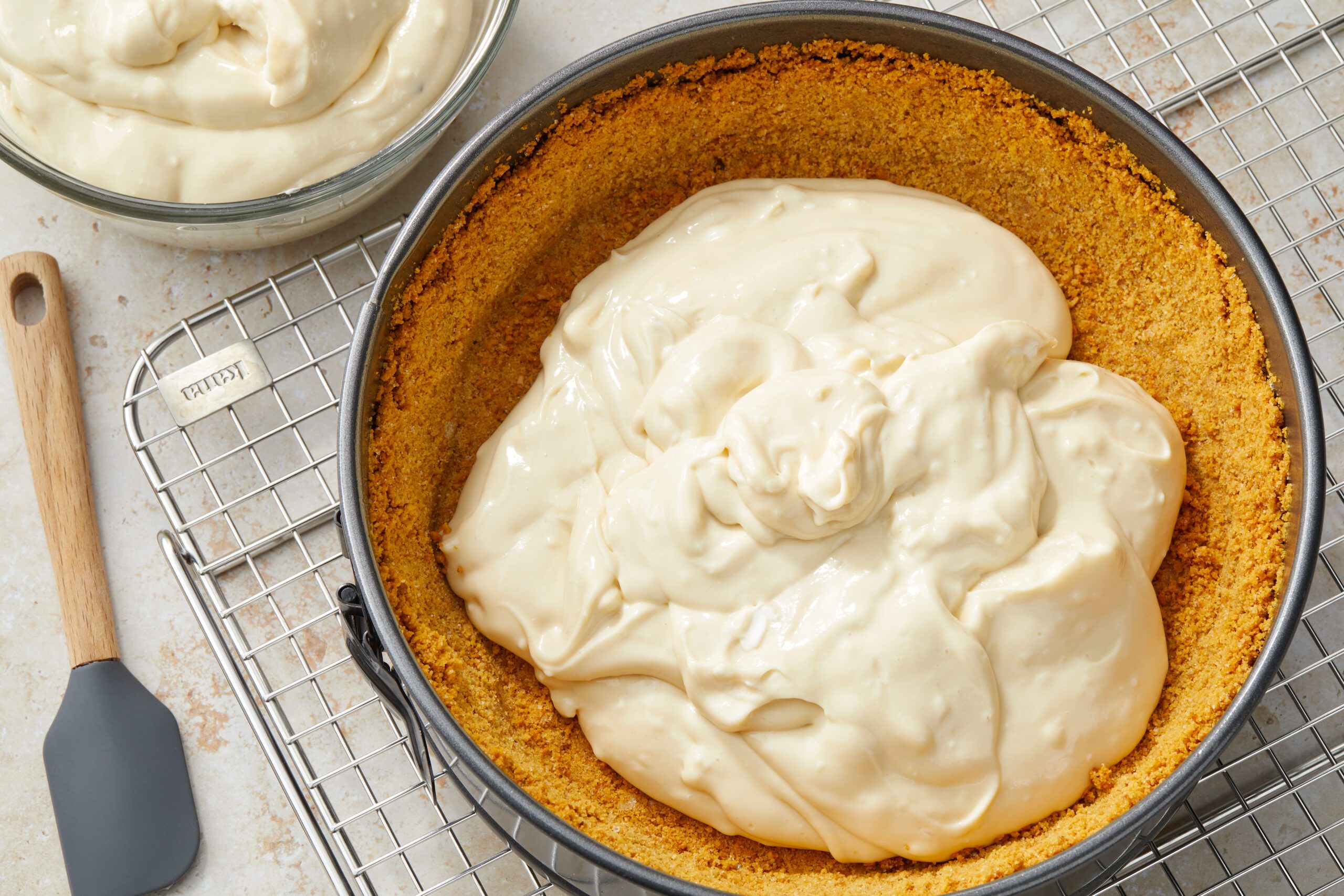 Cheesecake filling spread evenly in a graham cracker crust inside a round baking pan on a wire rack. A spatula rests beside the pan, and a bowl with more filling is in the background.