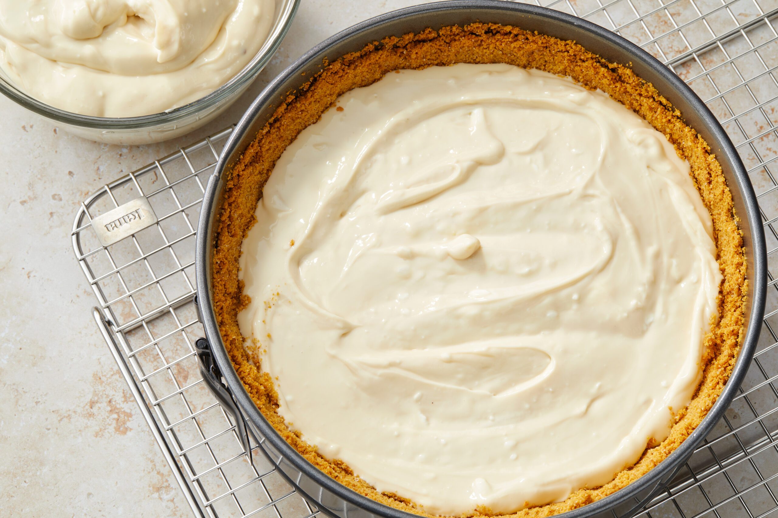 A cheesecake rests in a pan with a graham cracker crust, its creamy batter spread smoothly on top. It is set on a cooling rack next to a bowl containing more batter. The background is a light-colored countertop.