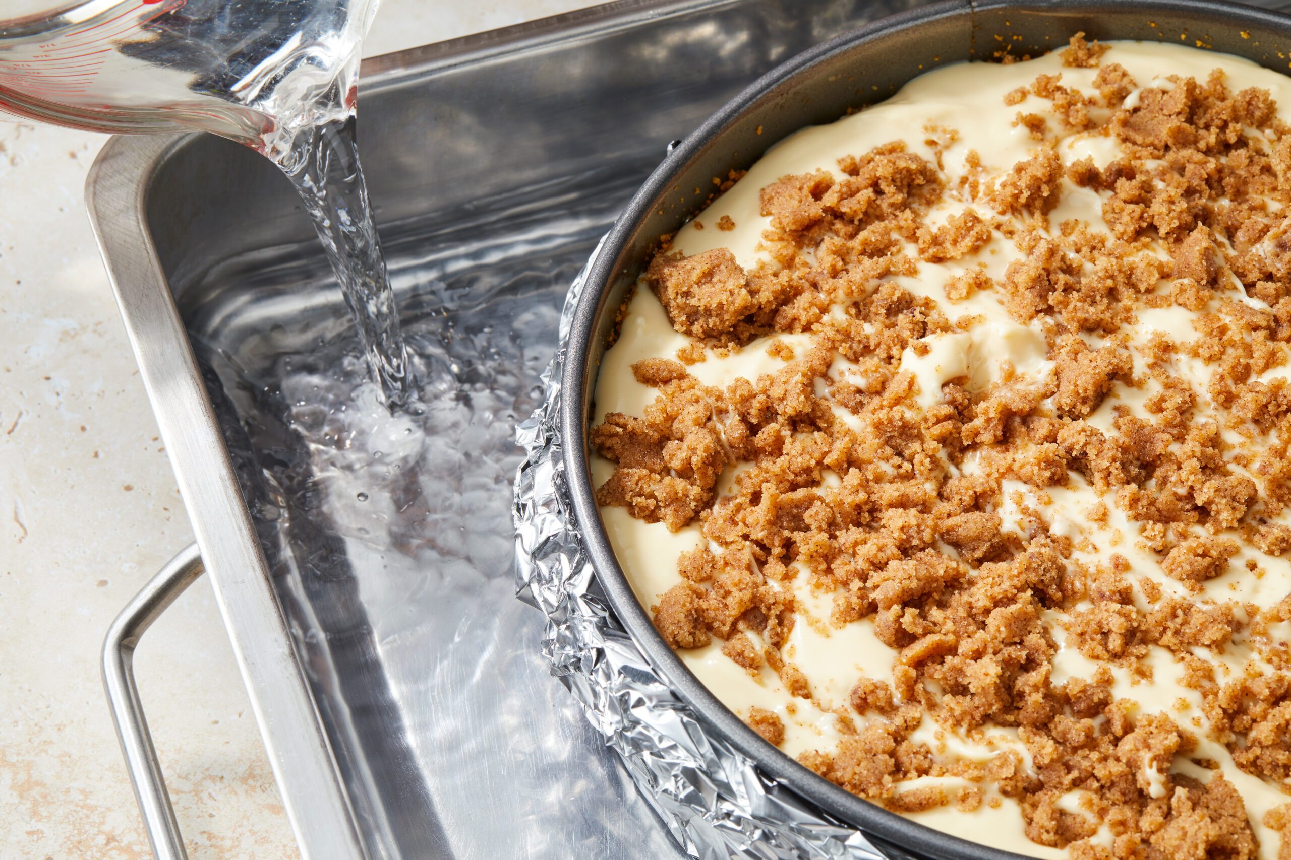 A cheesecake in a springform pan with a crumbled topping is wrapped in foil. It is placed in a larger pan, and water is being poured into the larger pan for a water bath before baking.