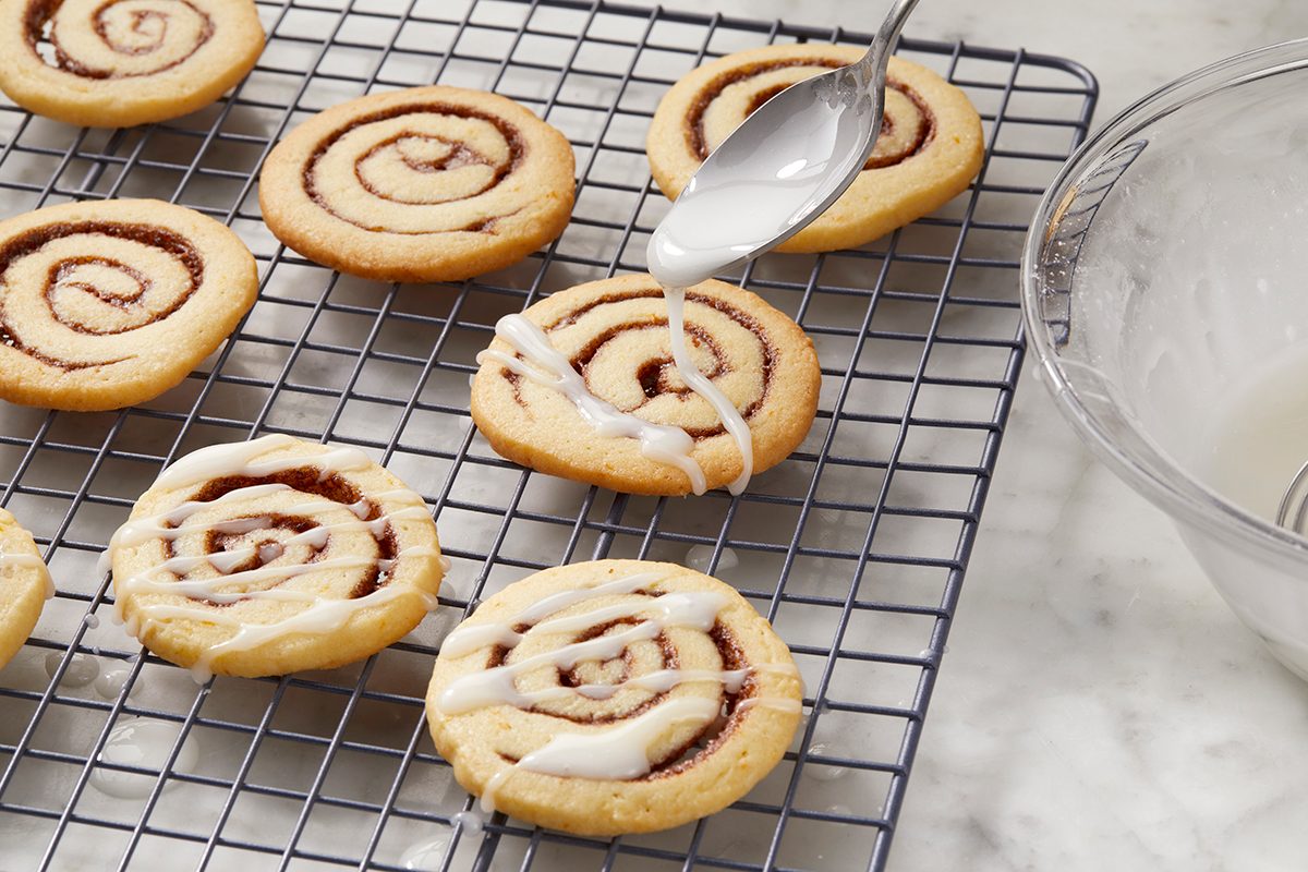 Cookies with spiral designs are on a cooling rack. A spoon is drizzling icing over one of them. A bowl with icing is nearby, on a marble countertop.