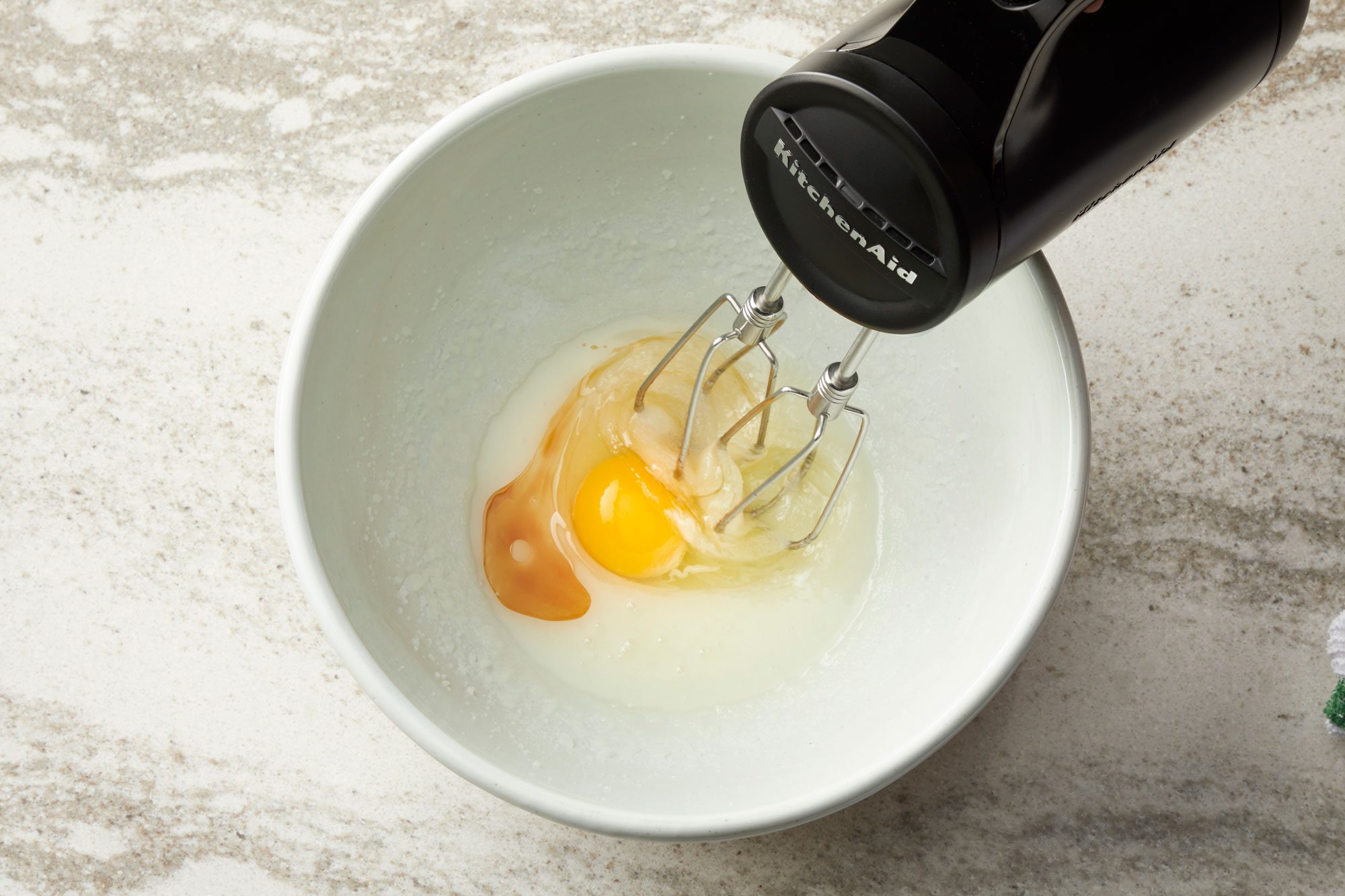 overhead shot of a white bowl sitting on a marble countertop; inside the bowl are the ingredients for a recipe; there are two eggs, one with a broken yolk spilling out, and a small amount of a clear or slightly yellowish liquid, possibly oil or melted butter; a black KitchenAid hand mixer is positioned with its beaters resting in the bowl