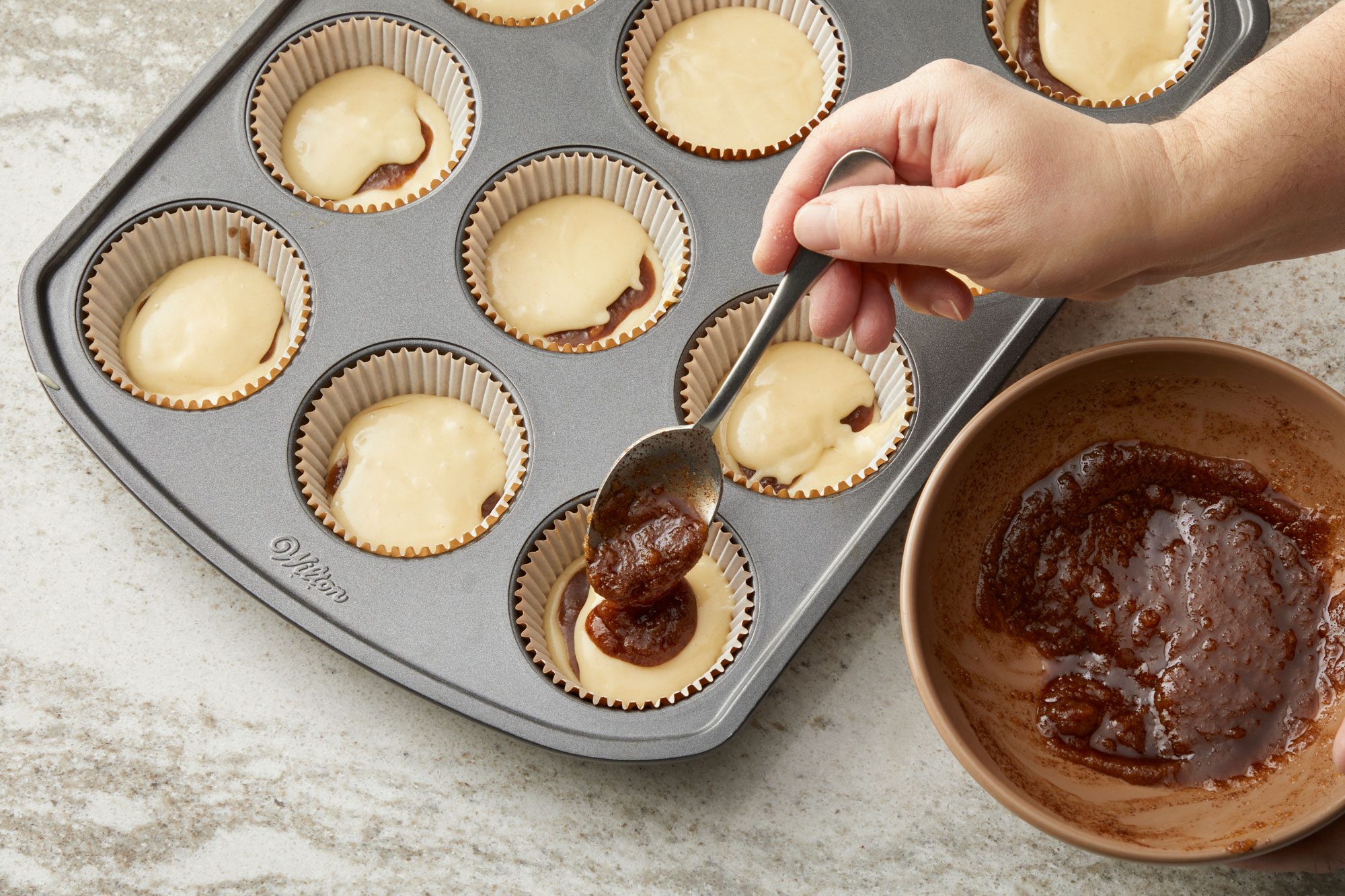 3/4th shot of a gray rectangular muffin tin filled with white paper liners and batter; several liners are already filled with light batter, while a few have a darker substance, likely a jam or fruit filling, added on top; a hand is visible in the right side of the frame, holding a silver spoon and adding more of the dark filling from a tan bowl; the bowl, partially visible, contains the thick, dark substance; the countertop is a light gray with a subtle speckled pattern