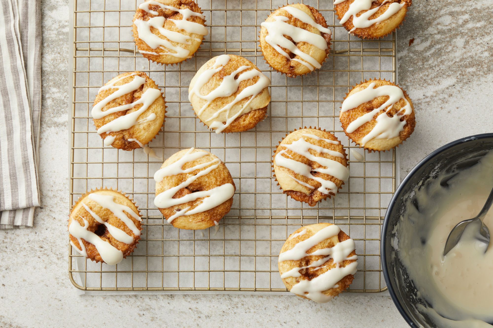 overhead shot of a batch of small, oval shaped baked cinnamon rolls, arranged on a gold wire cooling rack; drizzled with a white icing; in the lower right corner, a dark bowl containing leftover icing and a spoon is visible; the cooling rack sits on a light colored, speckled countertop