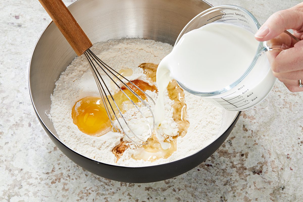 A person pours milk from a measuring cup into a mixing bowl containing flour, eggs, and vanilla extract. A whisk rests in the bowl on a marble countertop.