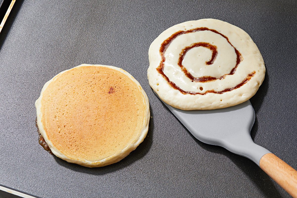 Two pancakes are cooking on a griddle. The left pancake is plain, while the right pancake has a cinnamon swirl pattern. A spatula is partially lifting the swirled pancake.