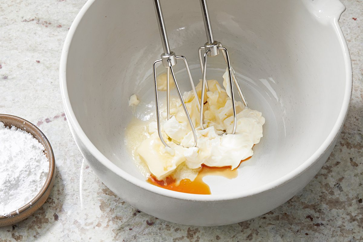 A white mixing bowl contains cream cheese, butter, and vanilla extract being blended with a hand mixer. Beside the bowl, there's a small dish of powdered sugar on a marble countertop.