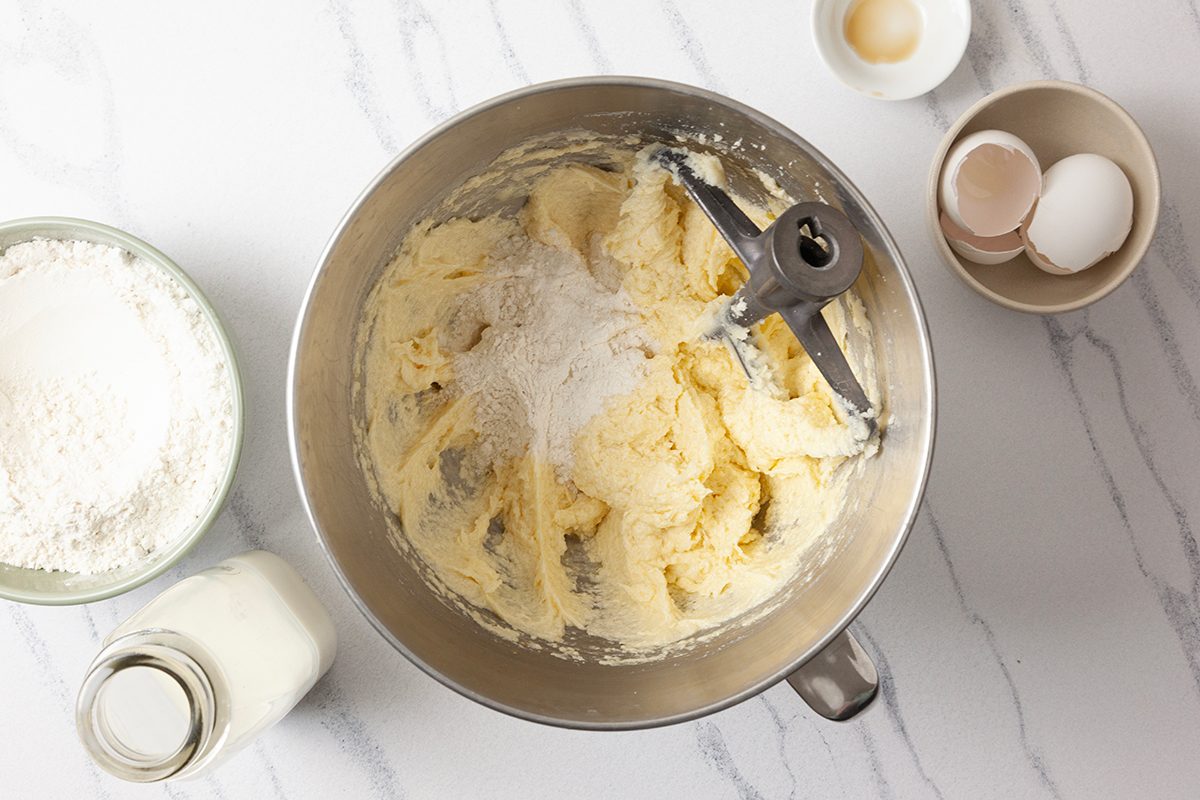 Mixing bowl with creamy batter and beater attachment on a white countertop. Nearby are bowls with eggshells, flour, and a bottle of milk, indicating preparation for baking.
