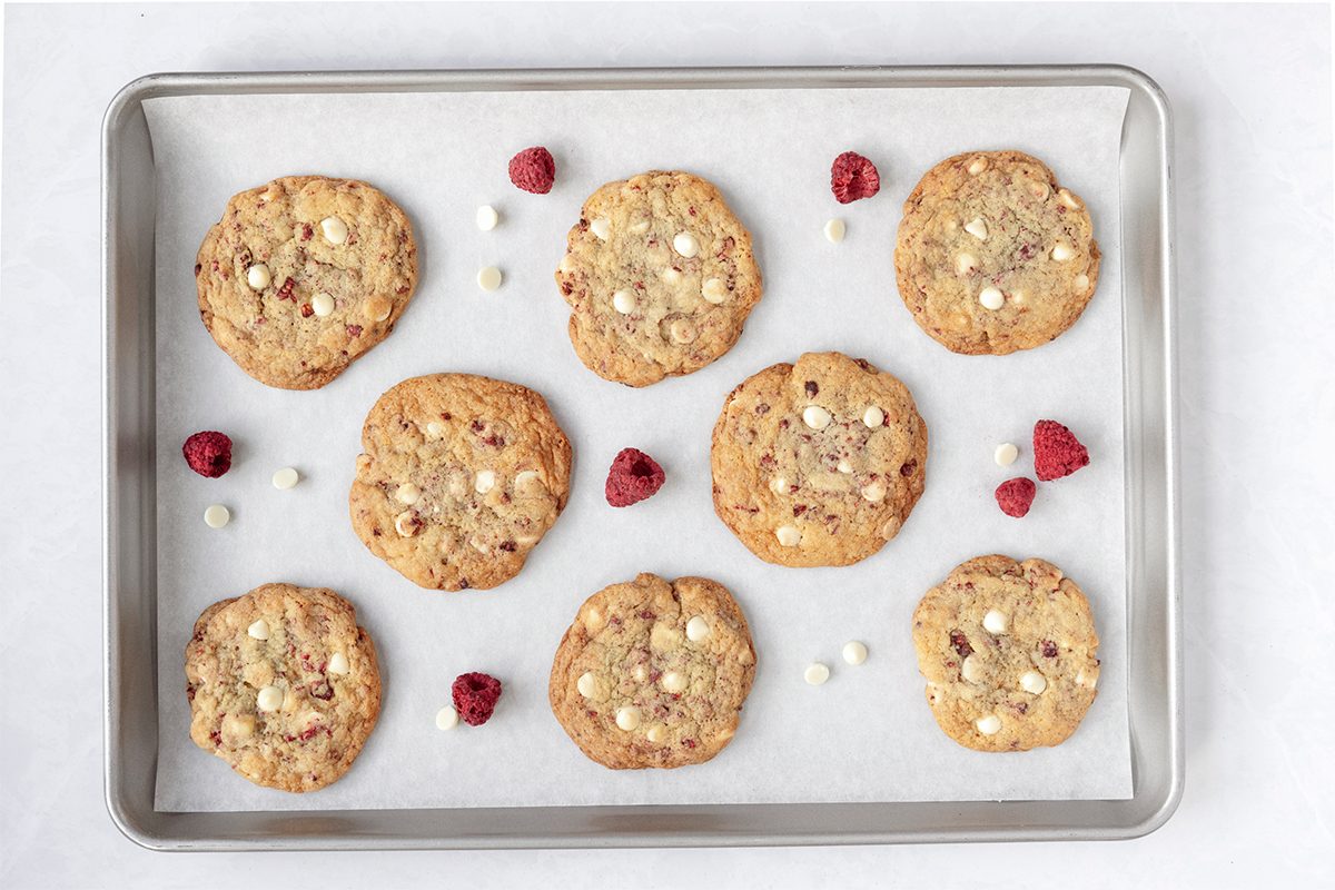 A baking tray with nine chocolate chip cookies and scattered raspberries. The cookies are arranged on parchment paper, showing a golden-brown color with visible white chocolate chips.