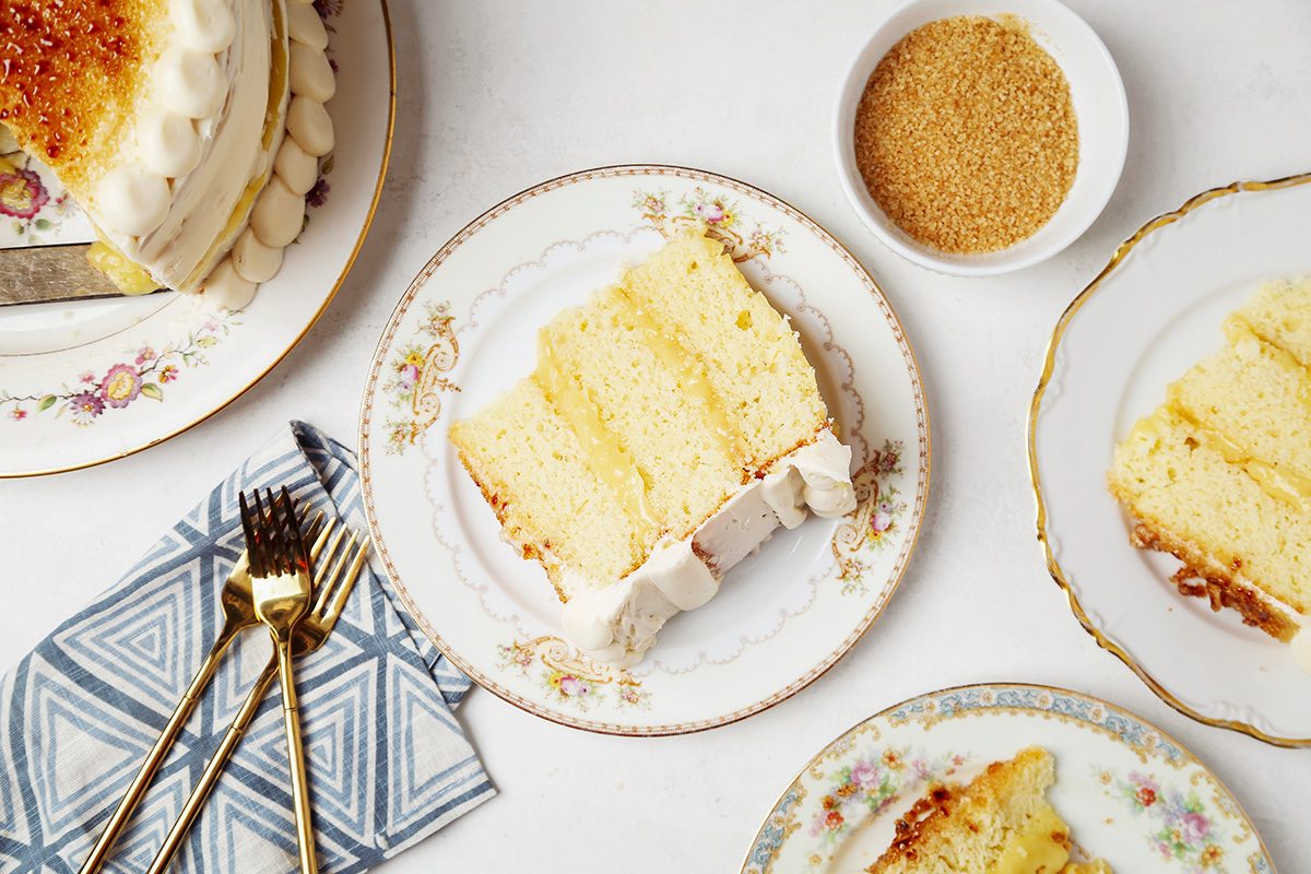 Slices of layered vanilla cake with frosting on elegant floral plates, accompanied by a bowl of brown sugar. A fork and knife rest on a patterned napkin nearby.