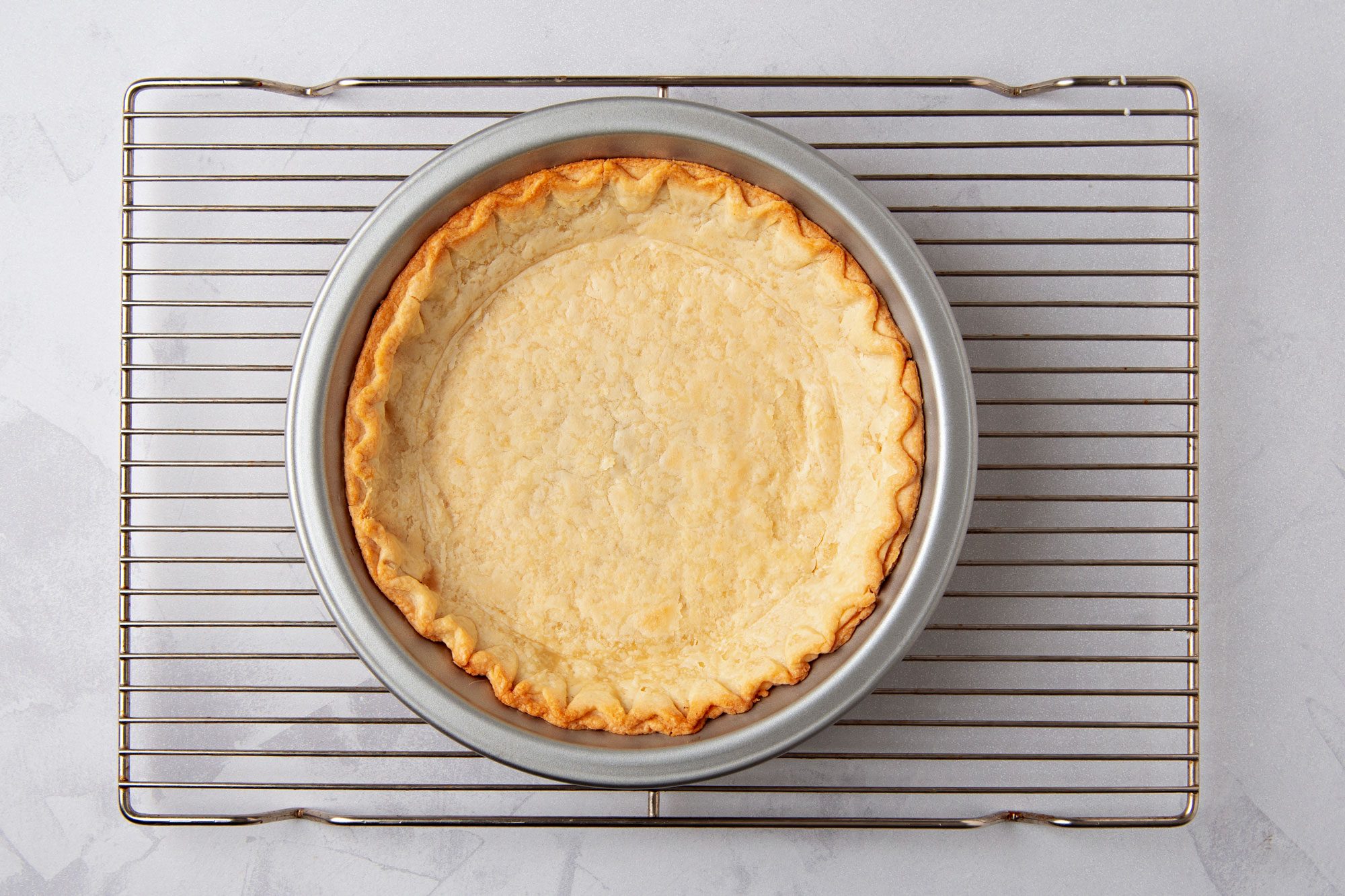 overhead shot of a pie crust in a pie pan on a silver cooling rack, against a white background; the pie crust is golden brown and has a crimped edge; the pie pan is a light silver color
