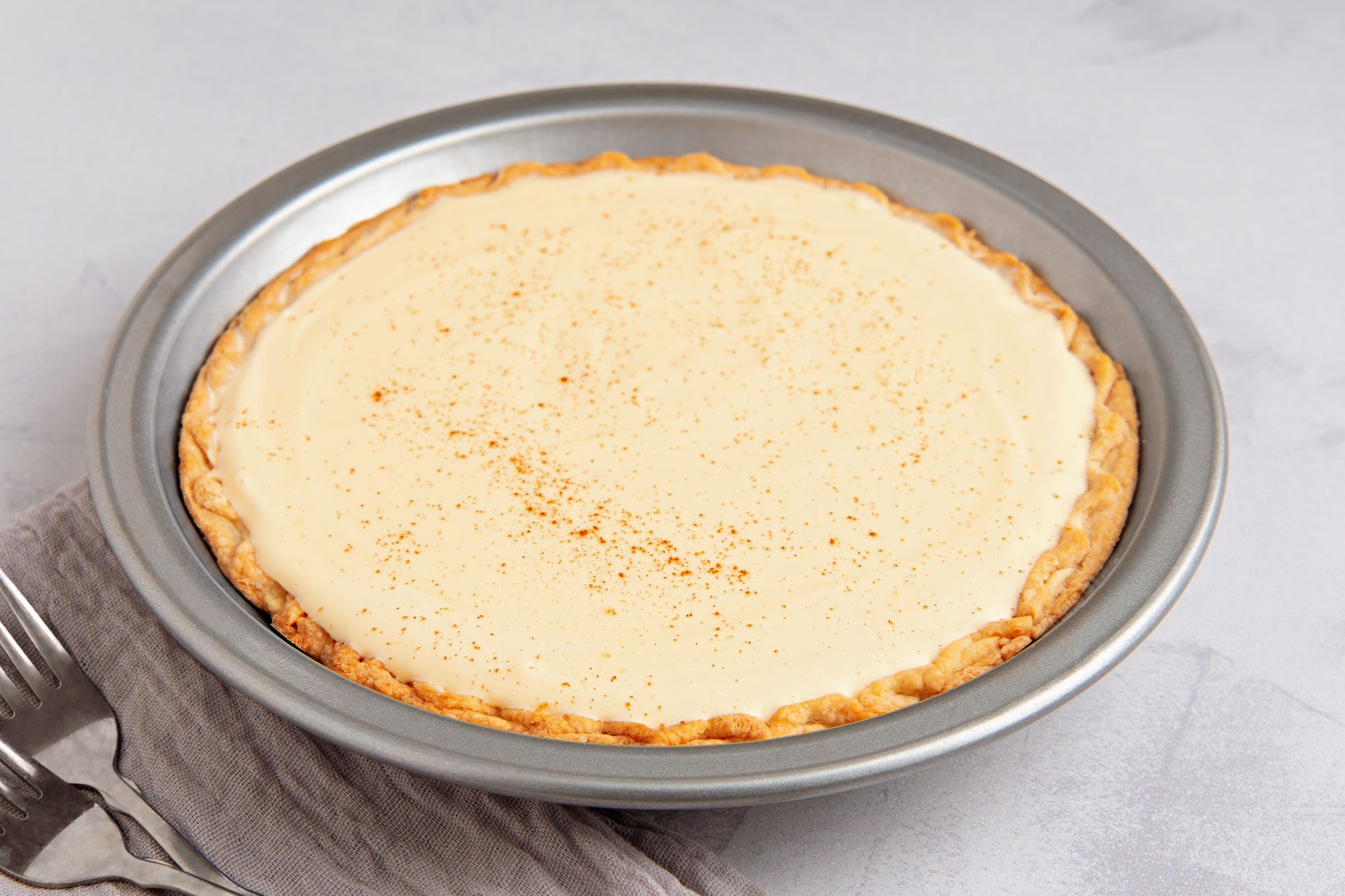 3/4th shot of a pie in a silver pie pan; the pie is filled with a light yellow custard-like filling, and it is sprinkled with a brown spice; the crust is golden brown; the pie is sitting on a white surface and there are two silver forks next to it on a grey napkin