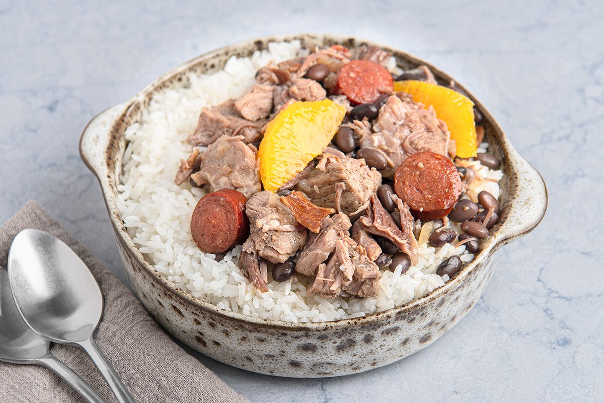 A bowl containing white rice topped with chunks of meat, sliced sausage, black beans, and orange segments.