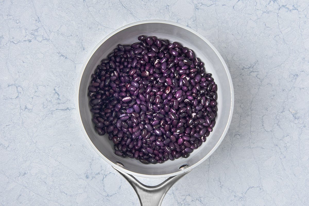 A silver saucepan filled with cooked black beans, placed on a light gray textured surface. The beans appear glossy and dark, contrasting with the metallic shine of the pan.