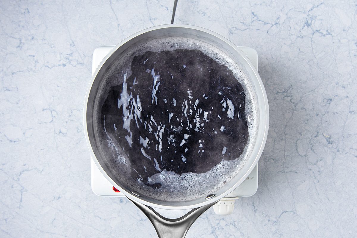 A stainless steel pot with boiling water on a white electric stovetop, viewed from above. The water appears dark, possibly infused with a dark ingredient, creating a rippling, bubbling effect over a light marbled surface.