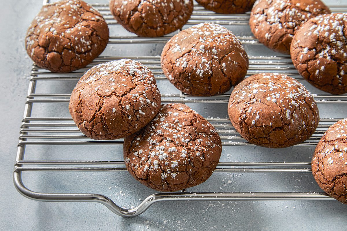 Chocolate crinkle cookies dusted with powdered sugar are cooling on a metal rack. The cookies have a cracked surface and are placed on a light-gray background.