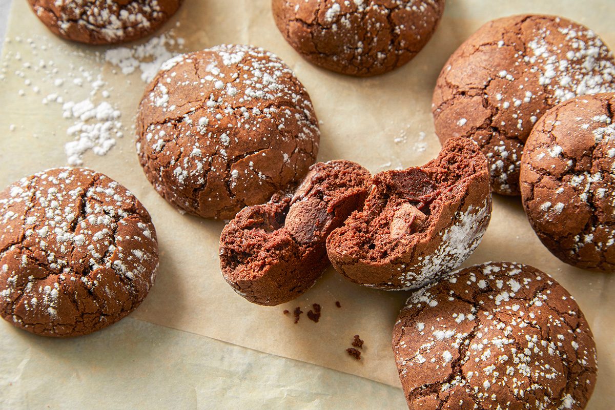 Chocolate crinkle cookies dusted with powdered sugar are arranged on parchment paper. One cookie is broken in half, revealing a soft, rich interior.