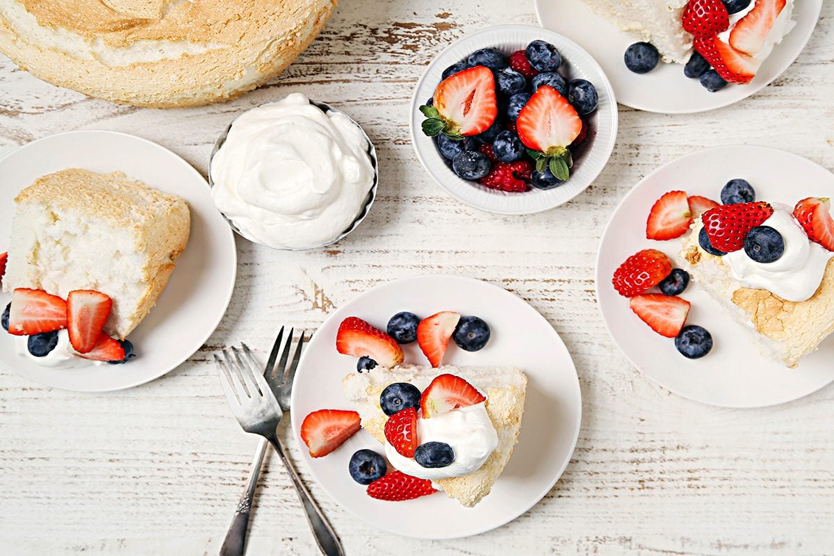Plates with angel food cake topped with whipped cream, strawberries, and blueberries on a white wooden table. A bowl of mixed berries and a bowl of whipped cream are nearby, along with two forks.