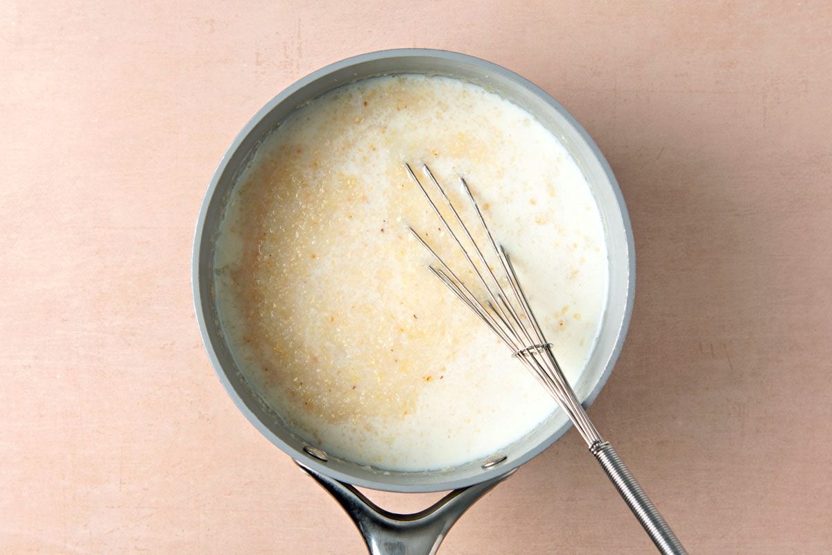 overhead shot of a pot filled with a white, creamy liquid and a whisk, on a light brown countertop; the creamy liquid is likely a milk-based sauce or a creamy soup; the whisk is partially submerged in the liquid